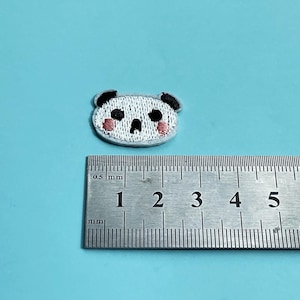 May include: Embroidered panda bear patch with black ears, eyes, and mouth, and pink cheeks. The patch is white with a gray border. A metal ruler is next to the patch, showing measurements in millimeters.