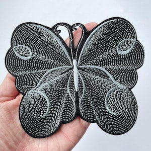 May include: An embroidered butterfly patch featuring a black base with intricate silver-grey stitching that forms the wing patterns and body details. The design includes circular accents on the wings and a central white vertical stitch line.