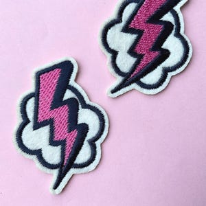 May include: Two embroidered patches featuring a pink lightning bolt design with black outlines, set against a white cloud shape. The patches have a black border and are displayed on a light pink background. These patches are suitable for clothing or accessories.