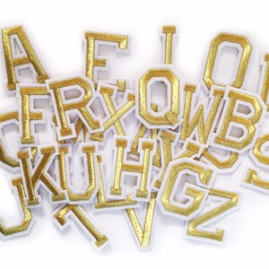 May include: A collection of varsity-style alphabet letter patches featuring a metallic gold finish with a white border. The letters are arranged in a scattered pile, showcasing various characters including A, E, I, O, D, F, R, Q, W, B, K, U, H, G, J, V, S, and Z.