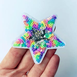 May include: A colourful star-shaped sequin patch, featuring a variety of colours such as pink, green, yellow, and purple. The sequins are attached to a light grey felt backing. The star is held in a hand.