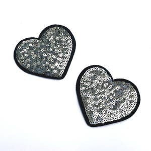 May include: Two heart-shaped patches. Each patch features silver sequins on a black felt backing. The sequins cover the entire heart shape, creating a shimmering effect. The patches are ideal for adding embellishment to clothing or accessories.