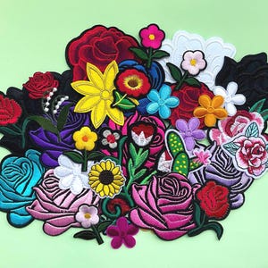May include: A selection of embroidered flower patches in diverse shapes and sizes. The patches feature roses, sunflowers, and other floral designs in red, pink, yellow, blue, and purple. The patches are arranged on a pale green background.