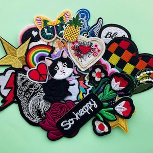 Może przedstawiać: Zbiór kolorowych naszywek do prasowania z różnymi wzorami, w tym tęczą, kotem, sercem, kwiatem, błyskawicą, gwiazdą, ananasem, sercem w kratkę i słowami "So happy".