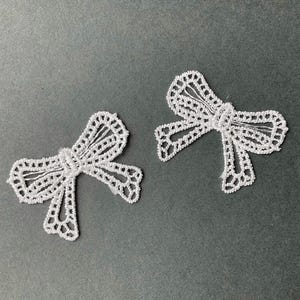 May include: Two white lace bow appliques are shown on a grey background. The bows have detailed openwork patterns, giving a delicate and airy look. These decorative elements are suitable for various craft projects and embellishments.
