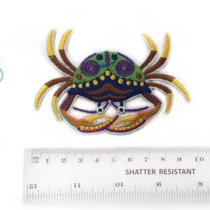 May include: A colorful embroidered iron-on patch of a crab with green, purple, yellow, orange, and brown details. The patch is approximately 5 cm wide.
