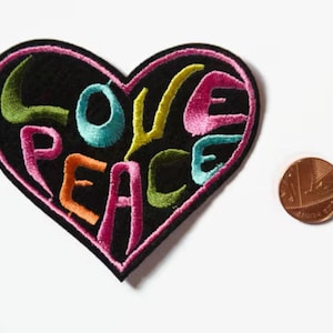 May include: A black heart-shaped patch with the words "LOVE PEACE" embroidered in colourful thread. The letters are in a stylised font and are outlined in pink.