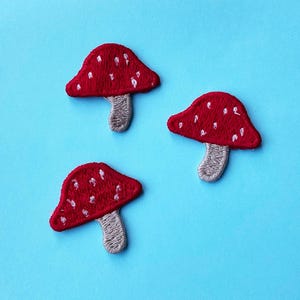 Op de afbeelding: Drie geborduurde patches van rode paddenstoelen met witte stippen en grijze stelen op een lichtblauwe achtergrond. De patches zijn gedetailleerd met gestikte contouren, wat een grillig, door de natuur geïnspireerd ontwerp oplevert. Geschikt voor kleding of accessoires.