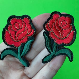 Op de afbeelding: Twee rode geborduurde rozenpatches met groene bladeren en gouden glitteraccenten. De patches zijn strijkbaar en kunnen worden gebruikt om kleding, tassen of andere items te versieren.