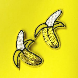 May include: Two yellow banana patches with black outlines and white embroidered tips. One banana patch is embellished with gold sequins. The patches are set against a bright yellow background.