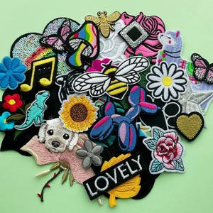 Op de afbeelding: Een verzameling geborduurde patches in verschillende vormen en kleuren, waaronder een bij, een vlinder, een bloem, een muzieknoot en een hond. Het woord "LOVELY" is geborduurd op een zwarte patch met een geel blad.