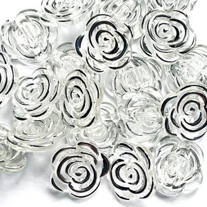 May include: A selection of clear, rose-shaped buttons with black detailing. These plastic buttons feature a floral design and are scattered on a white surface. Suitable for sewing and craft projects.