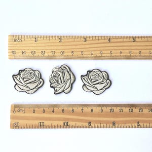 Op de afbeelding: Drie geborduurde rozenpatches met zwarte contouren en crèmekleurige centra worden getoond. Houten linialen met inch- en centimeter markeringen zijn boven en onder de patches geplaatst. De patches hebben een diameter van ongeveer 3.8 cm.