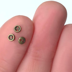 May include: Three small, antique brass-colored buttons with two holes each. The buttons are round with a slightly raised edge and are displayed on a person's finger.