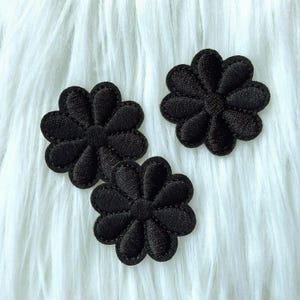 Può includere: Tre applicazioni di fiori in feltro nero con un design a margherita. Le applicazioni sono su uno sfondo bianco peloso.