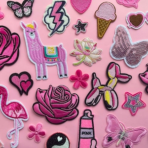 Op de afbeelding: Een verzameling roze geborduurde patches op een roze achtergrond. Patches zijn onder meer een lama, rozen, een flamingo, een vlinder, een bliksemschicht, een ijsje en het woord "PINK".