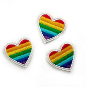 May include: Three embroidered heart patches with a rainbow design. Each heart features horizontal stripes in red, orange, yellow, green, blue, and purple, outlined with white stitching. The patches are on a white background.