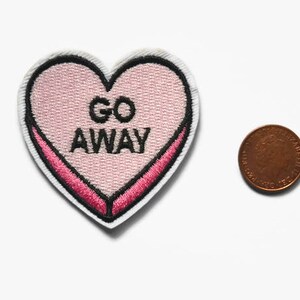 May include: Pink heart-shaped embroidered patch with the text "GO AWAY" in black lettering.