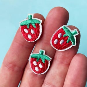 May include: Three embroidered strawberry patches. Each patch features a red strawberry with white seeds and a green stem. The patches are on a light blue background.