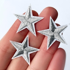 May include: Three silver star patches with white borders are displayed. The stars have a detailed embroidered texture, with a metallic sheen. These decorative patches can be used for clothing or craft projects.