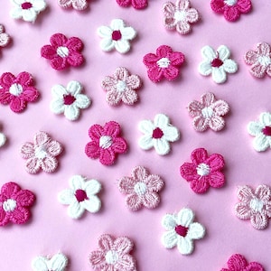 Könnte beinhalten: Eine Ansammlung von gestickten Blumenapplikationen in Rosa- und Weißtönen auf rosa Hintergrund. Die Blumen haben ein fünfblättriges Design mit kontrastierendem Zentrum, ideal für Bastelarbeiten und Verzierungen.