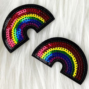 May include: Two rainbow-colored sequin patches. Each patch is a half-circle shape with a black border. The rainbow colors include red, orange, yellow, green, blue, and purple. The patches are on a white, fluffy surface.