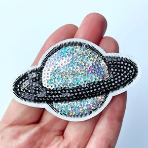 May include: A planet-shaped sequin patch with rings. The planet is filled with iridescent sequins, and the rings are black with black stitching. The patch is on a white background.