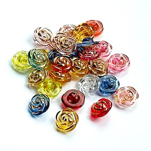 May include: A selection of rose-shaped buttons in a variety of colours, including red, yellow, blue, and clear. These translucent buttons are scattered on a white surface. Each button is approximately 1.5 cm in diameter.