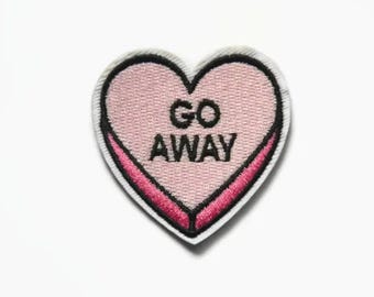 Op de afbeelding: Roze hartvormige patch met zwarte borduurwerk met de tekst "GO AWAY".