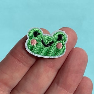 May include: Embroidered green frog patch with black eyes, a smiling mouth, and pink cheeks. The patch has a white border and is set against a light blue background. Ideal for adding to clothing or accessories.