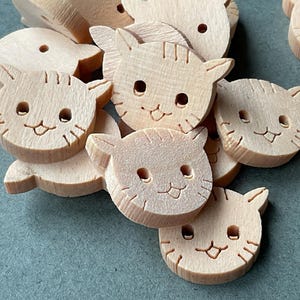 May include: A selection of unfinished wooden cat-shaped cutouts. Each cutout has a smiling face with etched details for the eyes, nose, and whiskers. The cat heads have pointed ears, ideal for craft projects.