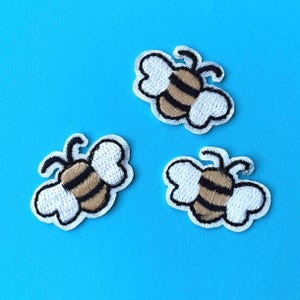 May include: Three embroidered bee patches are shown against a bright blue backdrop. Each patch features white wings, black antennae, and a brown and black striped body, all outlined in white.