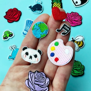 以下が含まれることがあります： さまざまな形と色の刺繍パッチのコレクション。パッチには、赤いバラ、緑の車、パンダ、地球、音符、パレット、蜂、ハート、陰陽のシンボル、パイナップル、クジラ、青い鳥が含まれています。