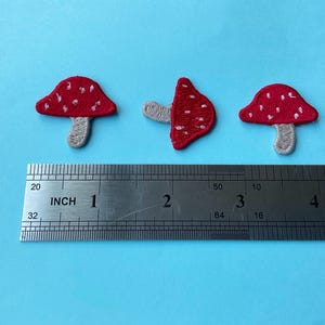 May include: Three embroidered mushroom patches with red caps featuring white spots and light beige stems. A metal ruler is positioned below the patches, showing they measure approximately 1 inch in width and height.