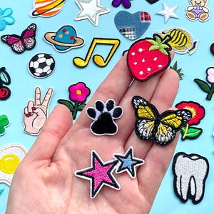 May include: A collection of colorful embroidered patches, including a strawberry, butterflies, a paw print, stars, and a musical note, on a light blue background. The patches are of various shapes and sizes, with detailed stitching.