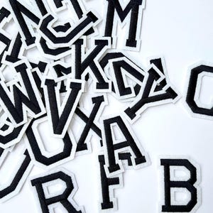 May include: A collection of black and white embroidered varsity letter patches. The letters are in a bold, block style with a black outline and white backing. The patches are scattered on a white surface.