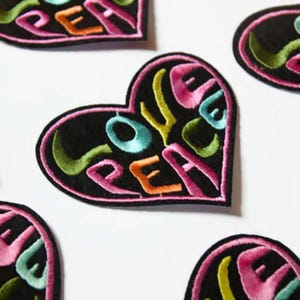 May include: A set of five black felt heart-shaped patches with the words "Love Peace" embroidered in various colours, including pink, green, blue, and orange.