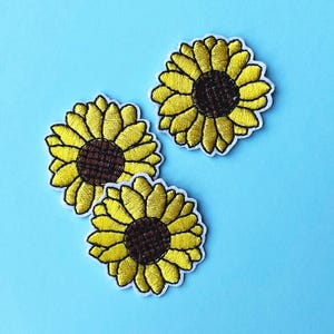 Puede incluir: Tres parches bordados de girasoles, con pétalos amarillos y centros marrones, están dispuestos sobre un fondo azul claro. Cada parche tiene un borde blanco. Son artículos de artesanía o costura.