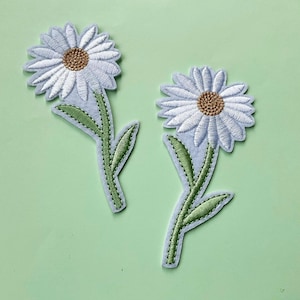 Puede incluir: Dos parches bordados de flores de margarita blancas con tallos y hojas verdes sobre un fondo verde claro.