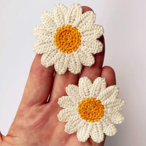 May include: Two handmade crochet daisy appliques. Each flower has white petals around a bright yellow centre. The appliques are held in a hand, showing the intricate stitch work and the contrast of colours.
