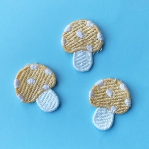 May include: Three embroidered mushroom patches with tan caps and white polka dots and stems, set against a light blue background. The patches are made of textured thread, and the design is simple and whimsical.