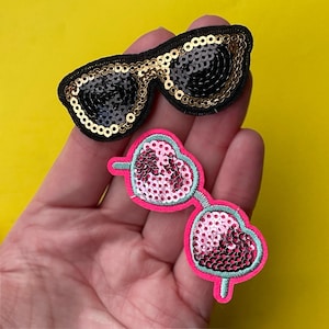 May include: Two sequined patches in the shape of sunglasses. One pair is black with gold trim, the other is heart-shaped with pink sequins and a neon pink border. The background is yellow.