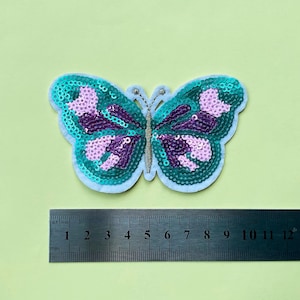 May include: A turquoise and purple sequined butterfly applique with a white outline. The butterfly is approximately 6 cm wide.