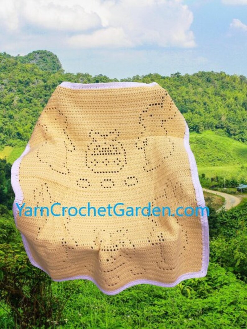 May include: A crocheted baby blanket in a light beige color with a white border. The blanket features a simple design of a bear with a heart in its paw. The blanket is displayed against a green background.