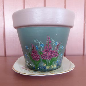 May include: Hand-painted flower pot with a sage green base and a light purple rim. The pot features a floral design with pink, blue, and yellow flowers. It sits on a white saucer with a gold rim, ideal for indoor or outdoor use.