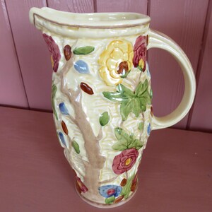 May include: A cream-coloured ceramic jug with a floral design. The jug features a tree motif with colourful flowers, leaves, and accents in red, yellow, blue, and brown. It has a curved handle and a spout for pouring.
