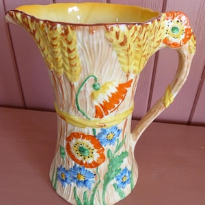 May include: A ceramic pitcher with a wheat and floral design. The pitcher has a yellow interior, a tan exterior with wheat stalks, and colourful flowers in orange, blue, and white. The handle is decorated with a flower.