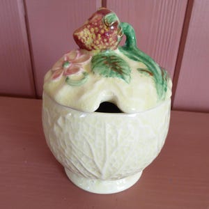 May include: A vintage ceramic sugar bowl with a lid, featuring a strawberry and floral design. The bowl is light yellow with a leaf pattern, and the lid has a strawberry-shaped handle. The overall design is charming and decorative.