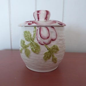 May include: A white ceramic jar with a lid, decorated with a floral design. The jar has a textured surface and is adorned with pink rose blossoms and green leaves. The lid features a matching rose design.