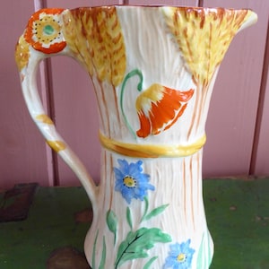 May include: A cream-coloured ceramic pitcher decorated with wheat, orange poppies, and blue flowers. The handle features a poppy design. A yellow band encircles the pitcher's midsection. The pitcher has a spout for pouring.